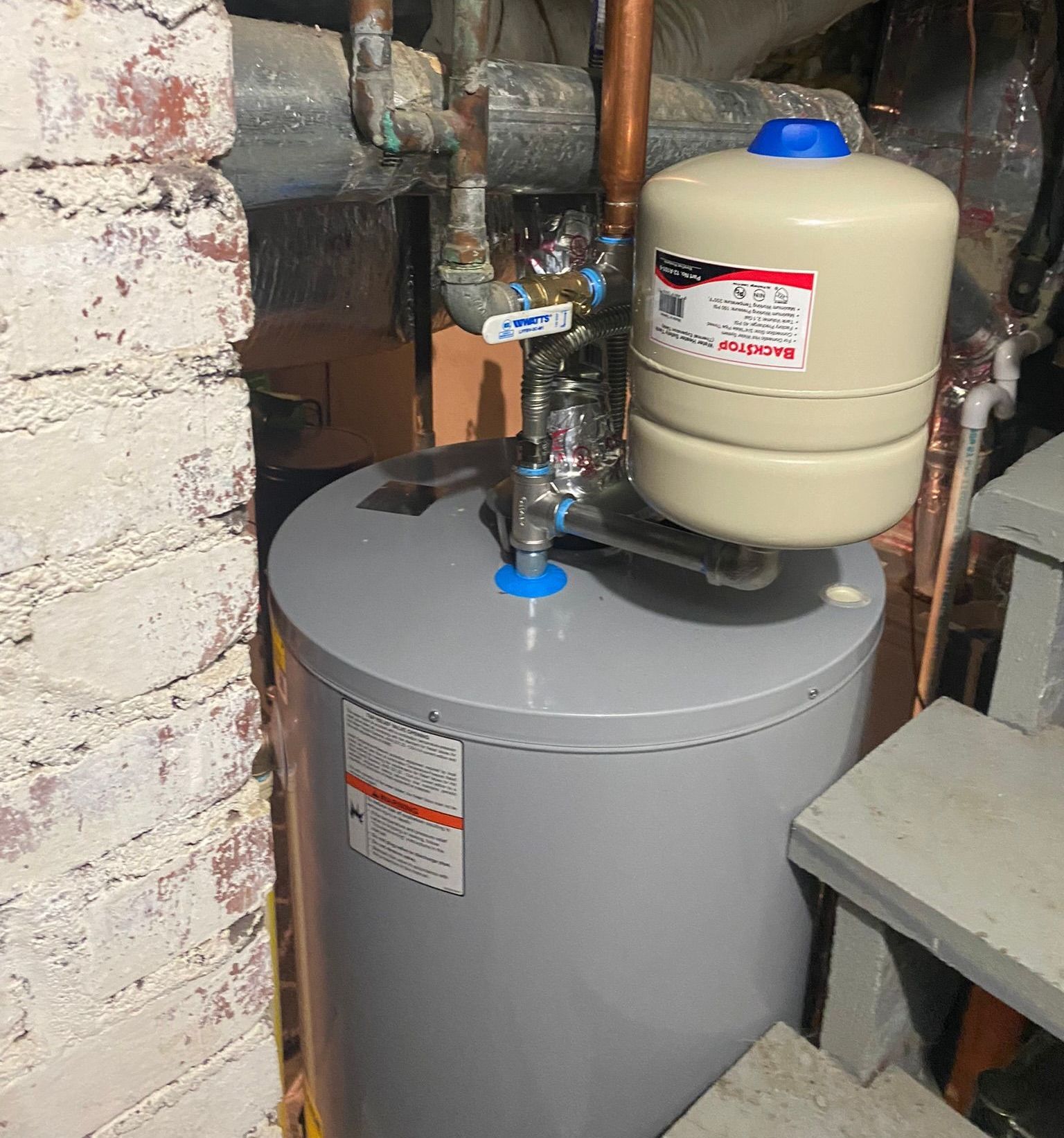 A water heater is sitting in a basement next to a brick wall.