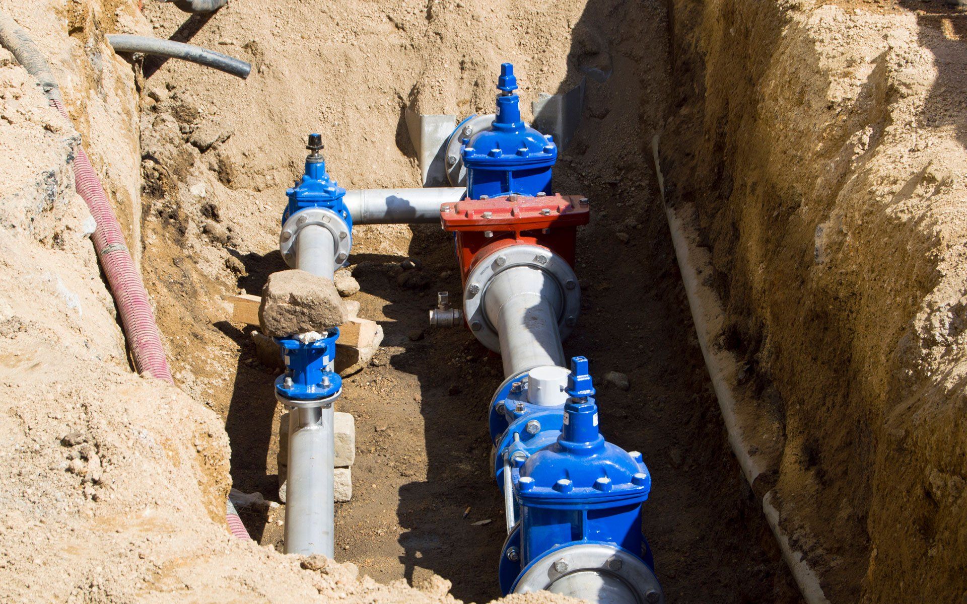 Pipes with blue and red valves in a trench filled with sand and dirt.