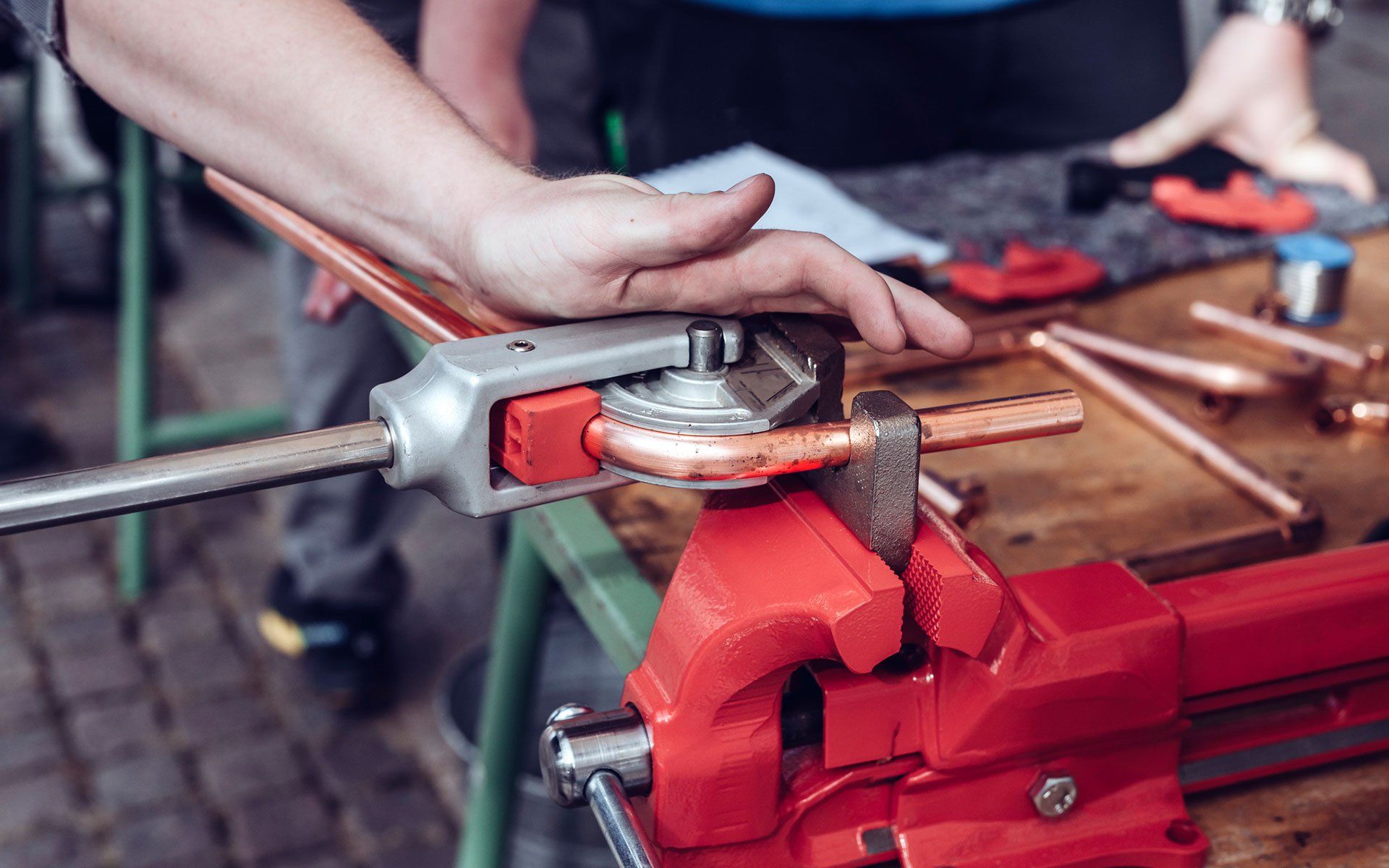 A person uses a threader to cut threads on a copper pipe held in a red vise on a wooden table.