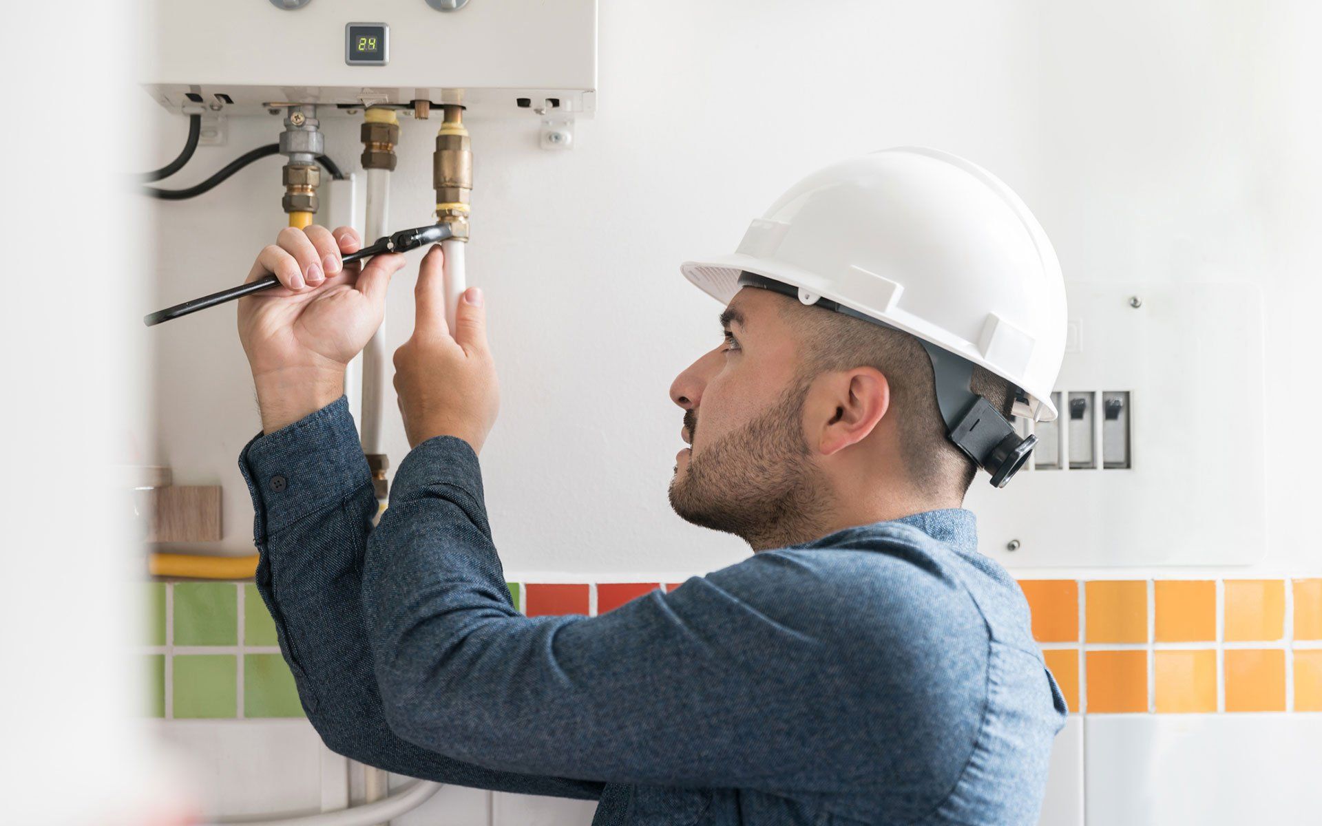 A man in a hard hat repairs plumbing, using a wrench, near a white appliance and orange tiles.