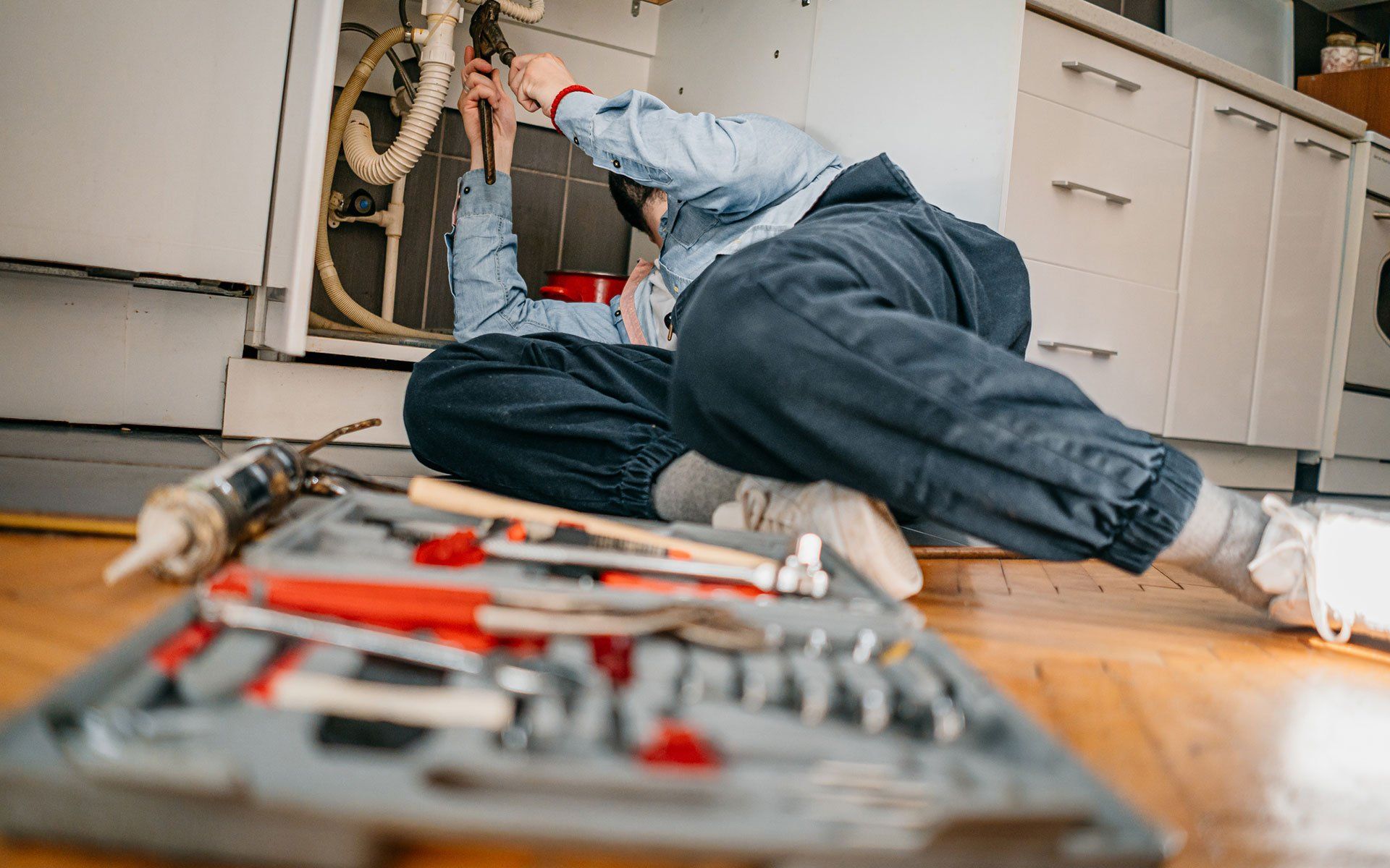 Person in blue overalls under kitchen sink fixing pipes, tools in foreground.