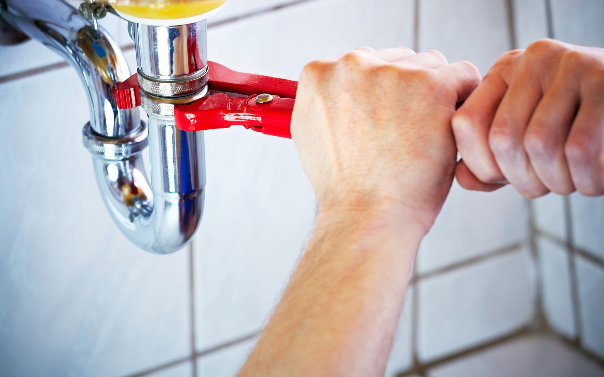 Hands using a red wrench to tighten a chrome pipe under a sink.