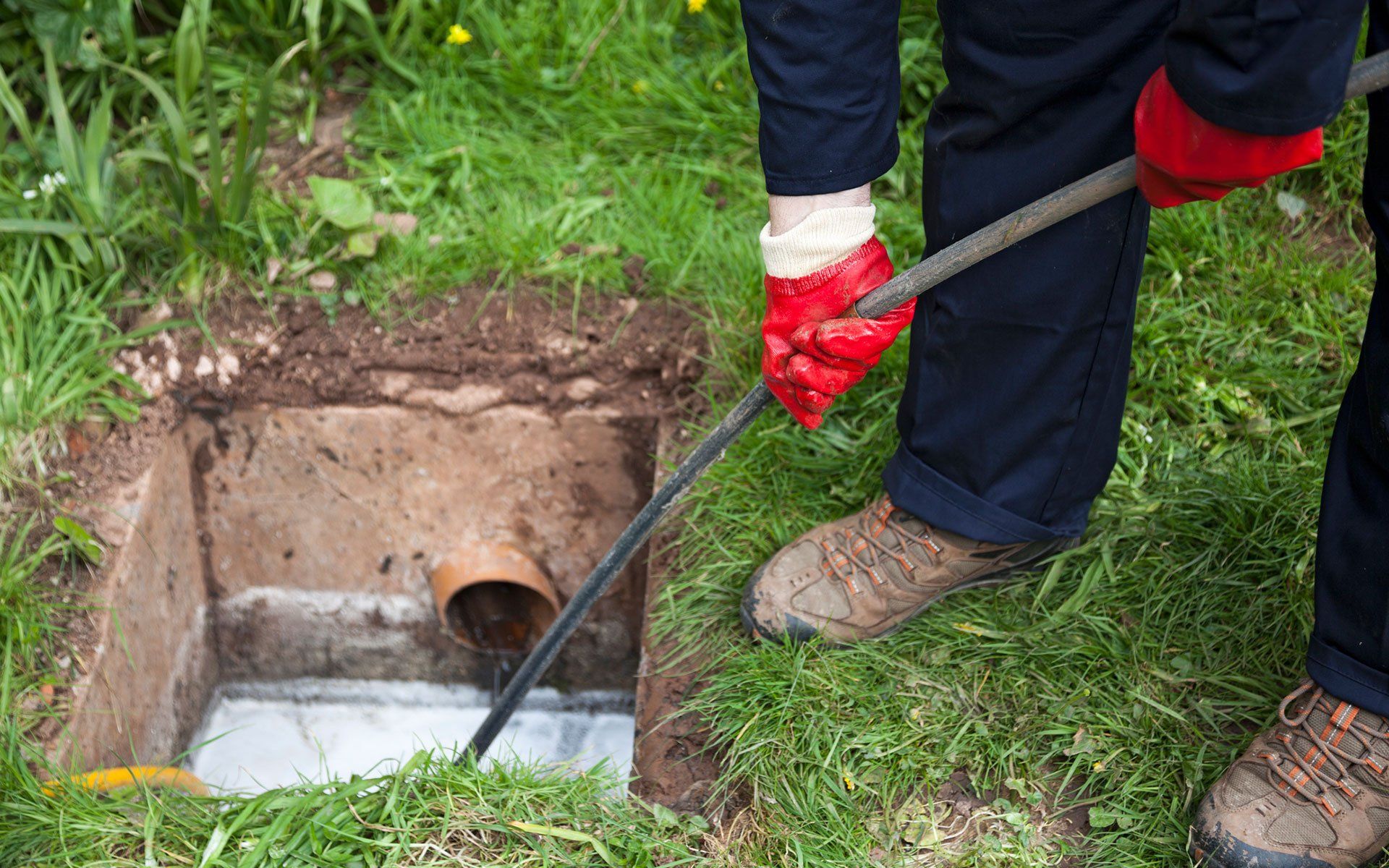 Person in red gloves using a drain snake to clear a pipe in a yard.