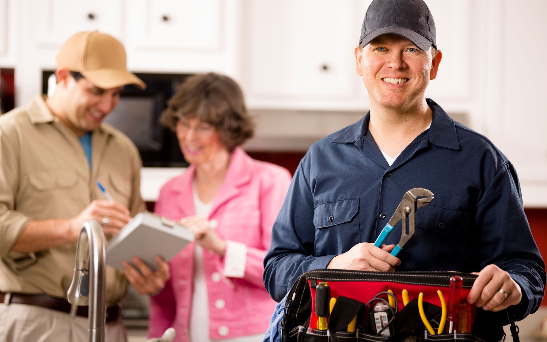 Plumber with tool bag smiling, standing in kitchen with two people signing a contract.