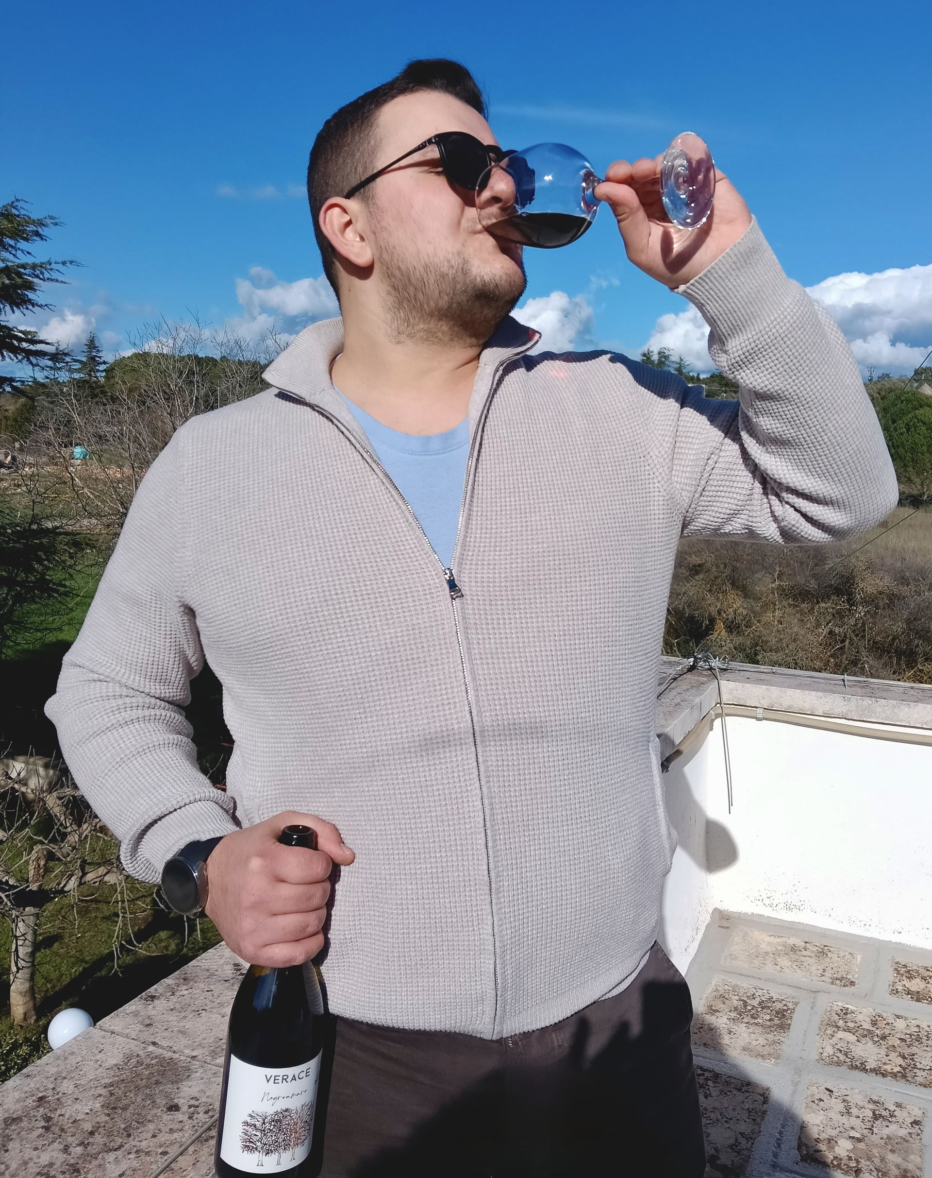 Uomo che beve vino rosso all'aperto, con in mano una bottiglia. Sfondo cielo azzurro.