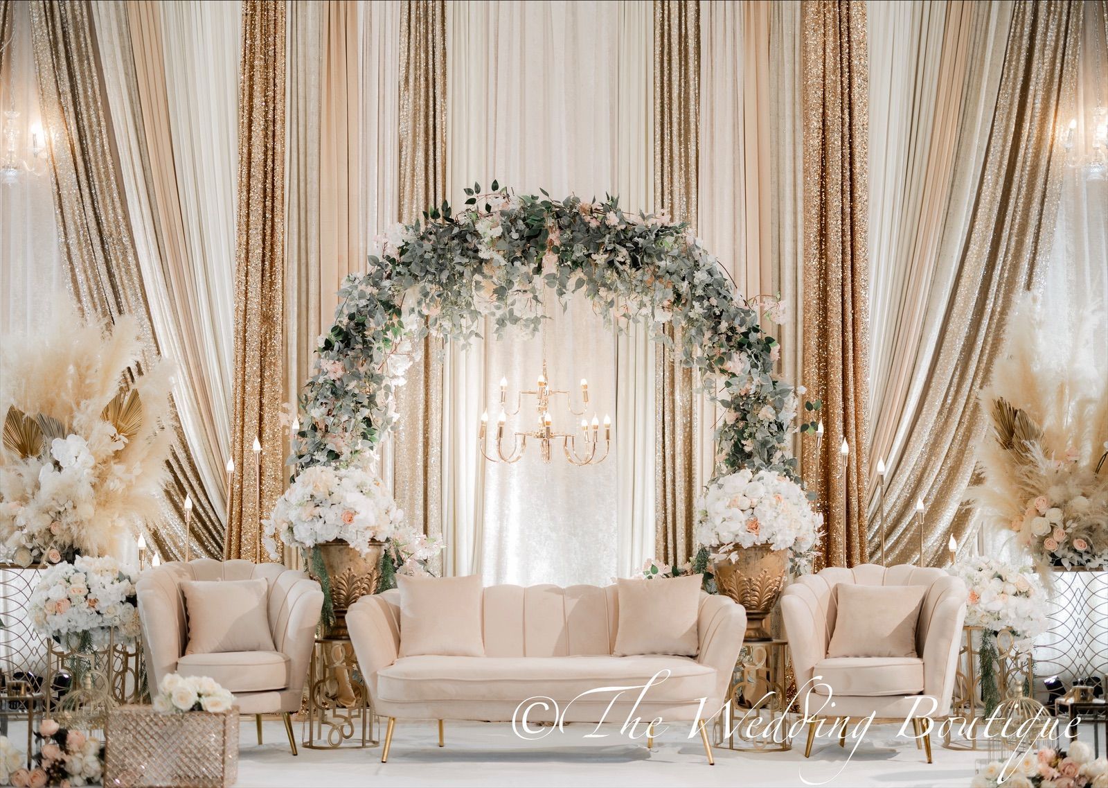 An arch of flowers going over a white sofa and chairs with gold and white curtains on the backdrop