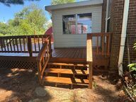 Wooden deck with stairs, leading to a room with windows, next to a brick wall.