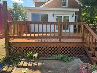 Wooden deck with lattice base, in front of a white-walled house, brown railing, green plants below.