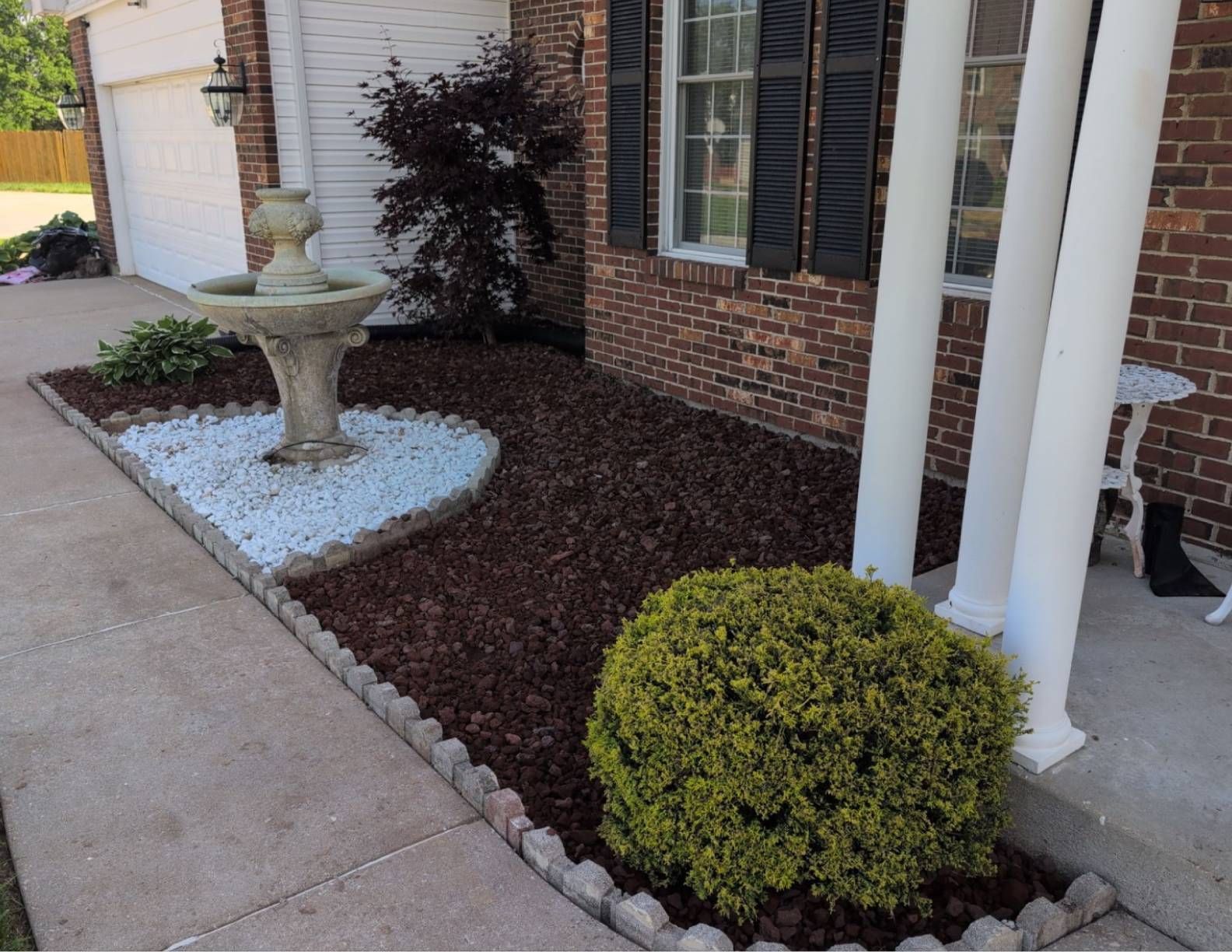 A front yard with a fountain, dark mulch, and a green bush next to a brick house.