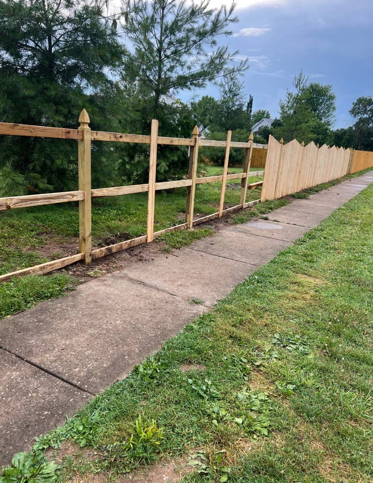 Wooden fence and sidewalk bordering grass, trees and cloudy sky.