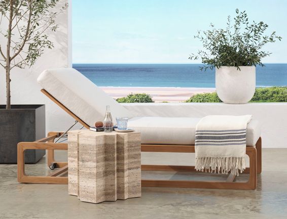 A lounge chair with a blanket on it and a side table in front of a window overlooking the ocean.