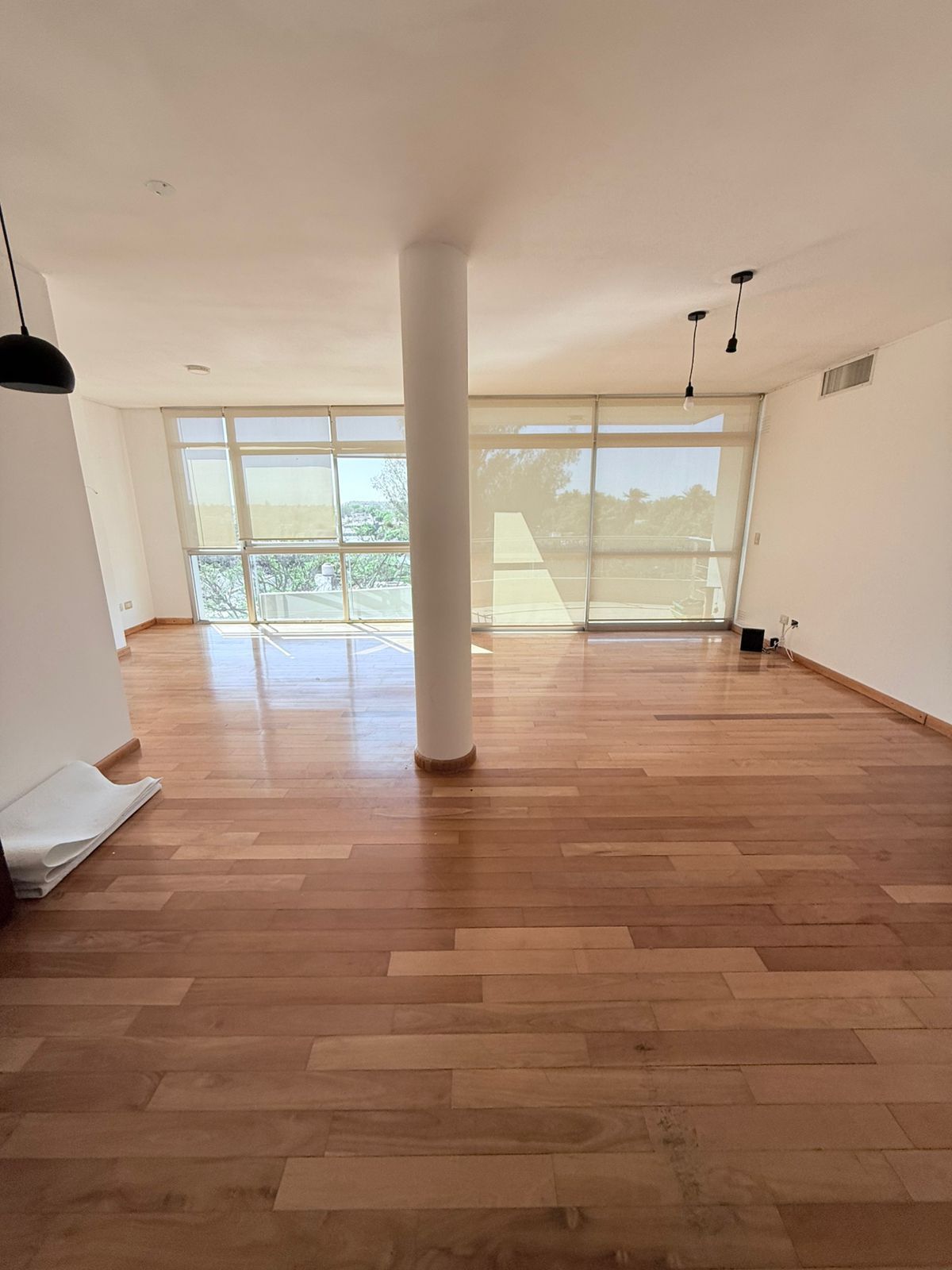 Habitación vacía con pisos de madera, ventanas grandes y un pilar central blanco.