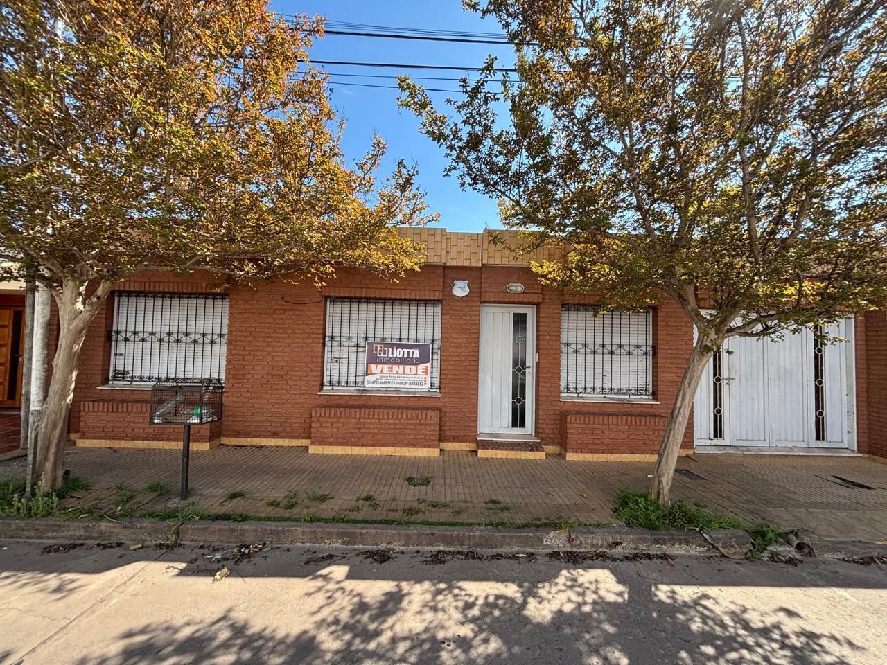 Edificio de ladrillo rojo con ventanas enrejadas, cartel de venta y árboles.