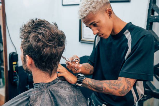 Barber cutting a client's hair with scissors in a salon. The barber has blonde hair and tattoos; the client has dark hair.