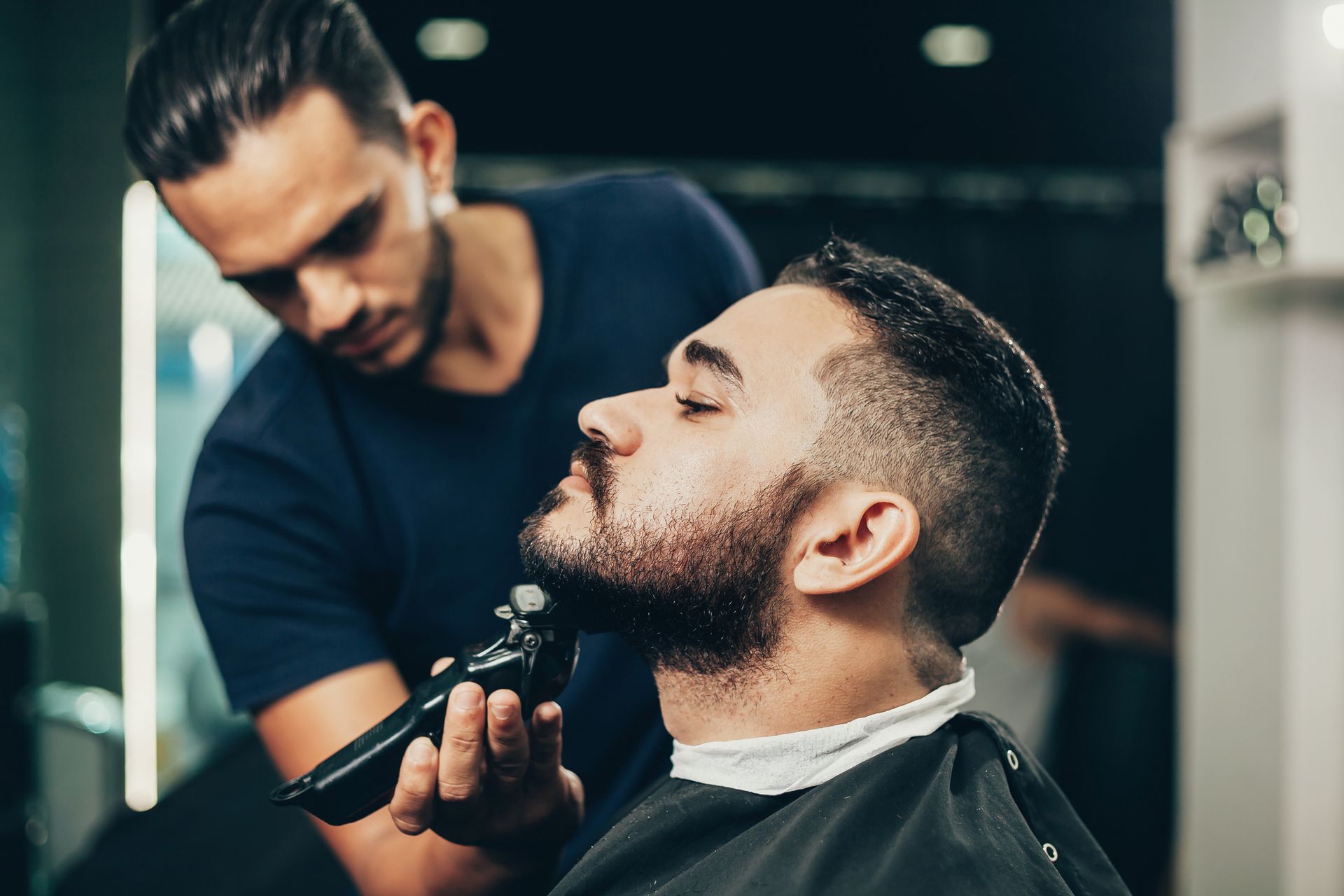 Barber trimming a customer's beard with a trimmer. The customer is in a barber chair.