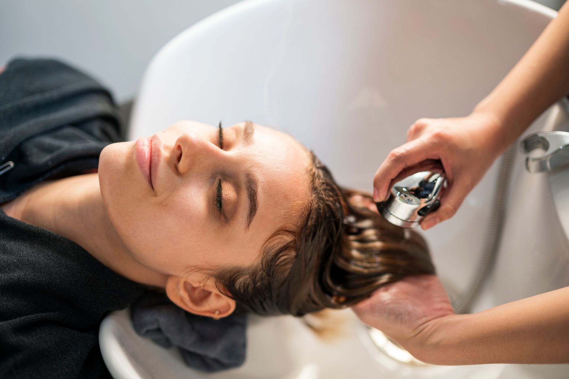 Woman having hair washed at a salon, eyes closed, hand holding a water sprayer over her hair.