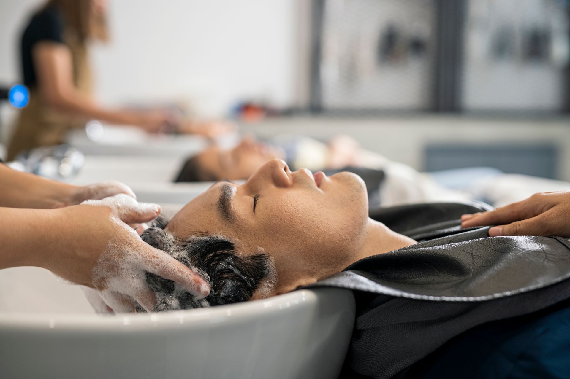Man having his hair washed at a salon, face relaxed. Hands in frame, white sink, person in background.