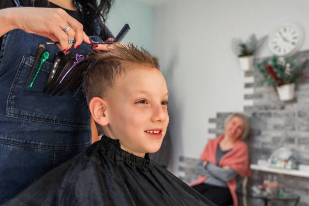 Boy getting a haircut at a salon, smiling. Barber holding scissors, tools in apron pocket. A woman watches in the background.