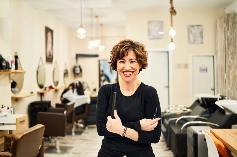 Woman smiling in a hair salon, holding scissors, standing near chairs and mirrors.