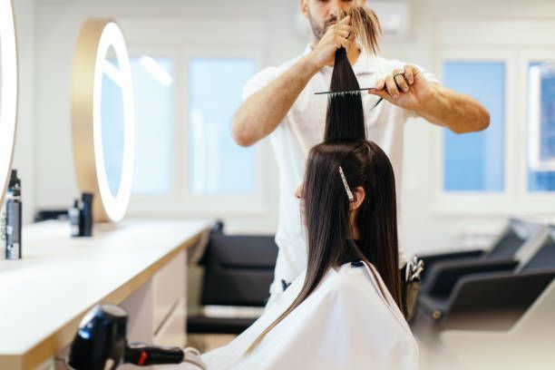 Hairdresser cutting long hair in a salon; woman in a cape sits, looking forward.