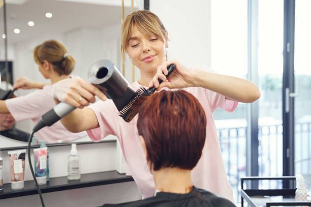 Woman blow-drying a client's hair in a salon. Client sits in chair.