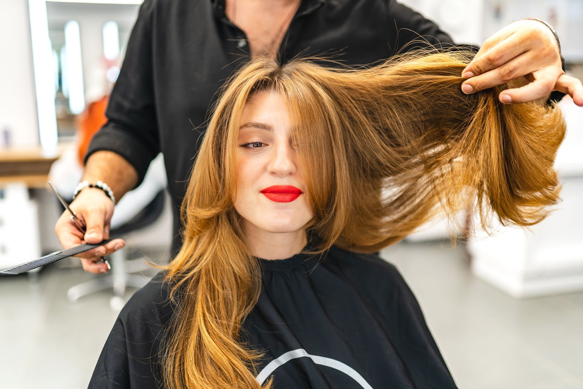 Woman in a salon with a stylist cutting her long, layered auburn hair; she wears red lipstick.