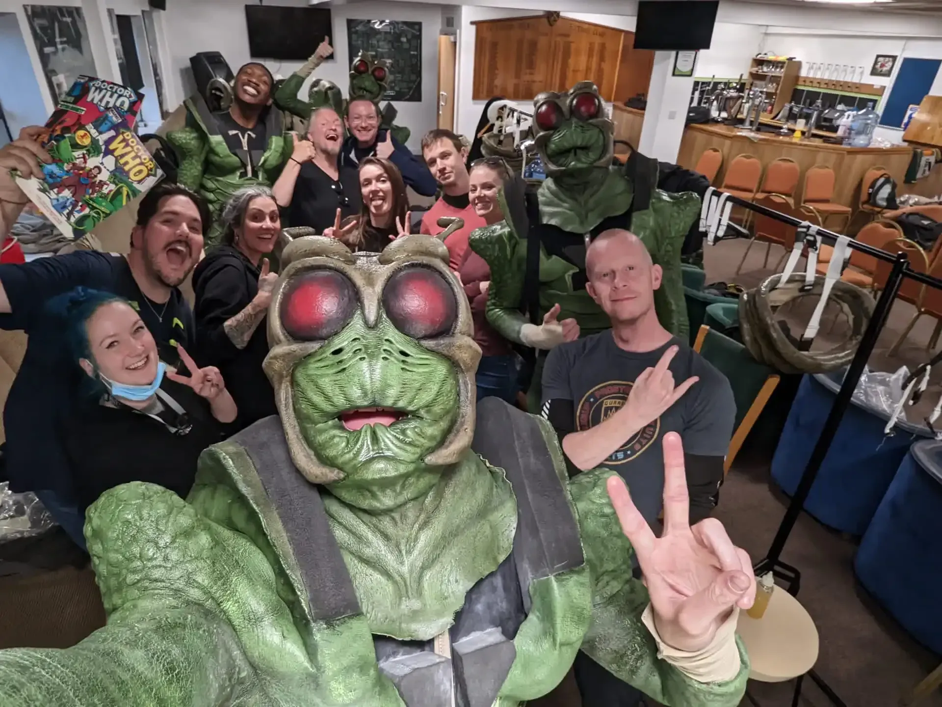 A group of people, including one in a green insect costume, posing for a selfie in a room with tables and chairs.