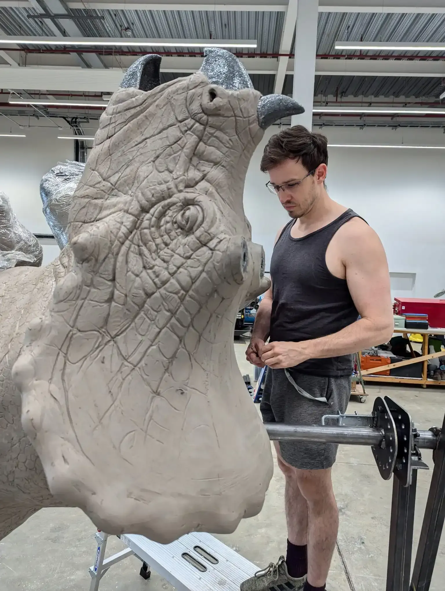 A person works on a large, textured clay sculpture of a horned dinosaur head in a studio.