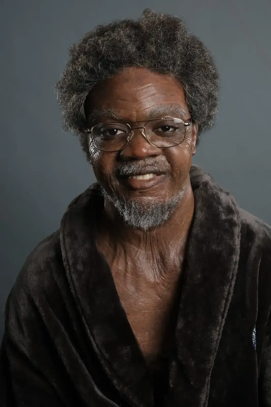 Man with glasses, gray afro, and beard smiles, wearing a dark brown robe, sweat visible on his skin.