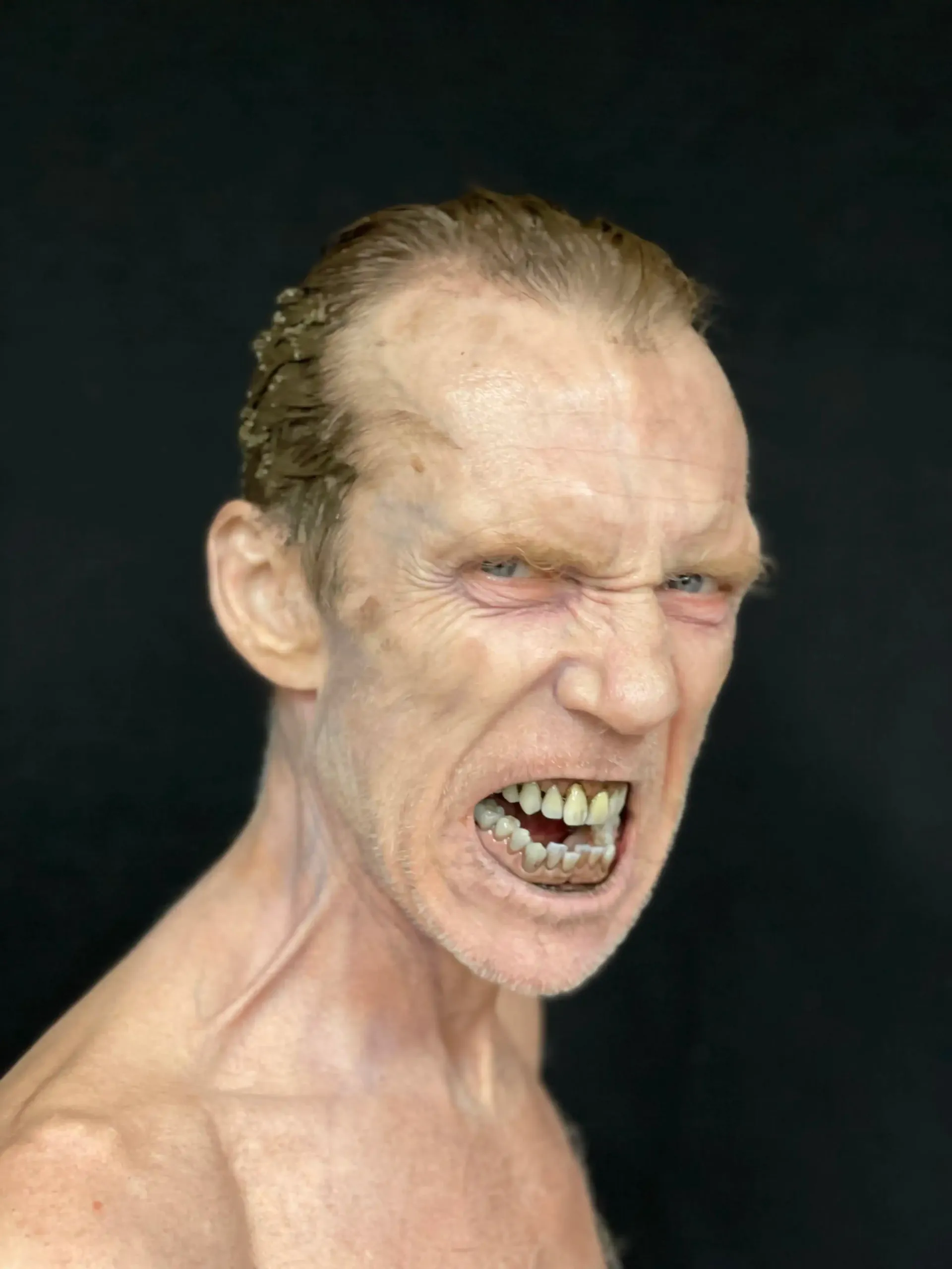 Frowning face with bared teeth, skin with fine lines, and short, slicked-back hair; against a black backdrop.