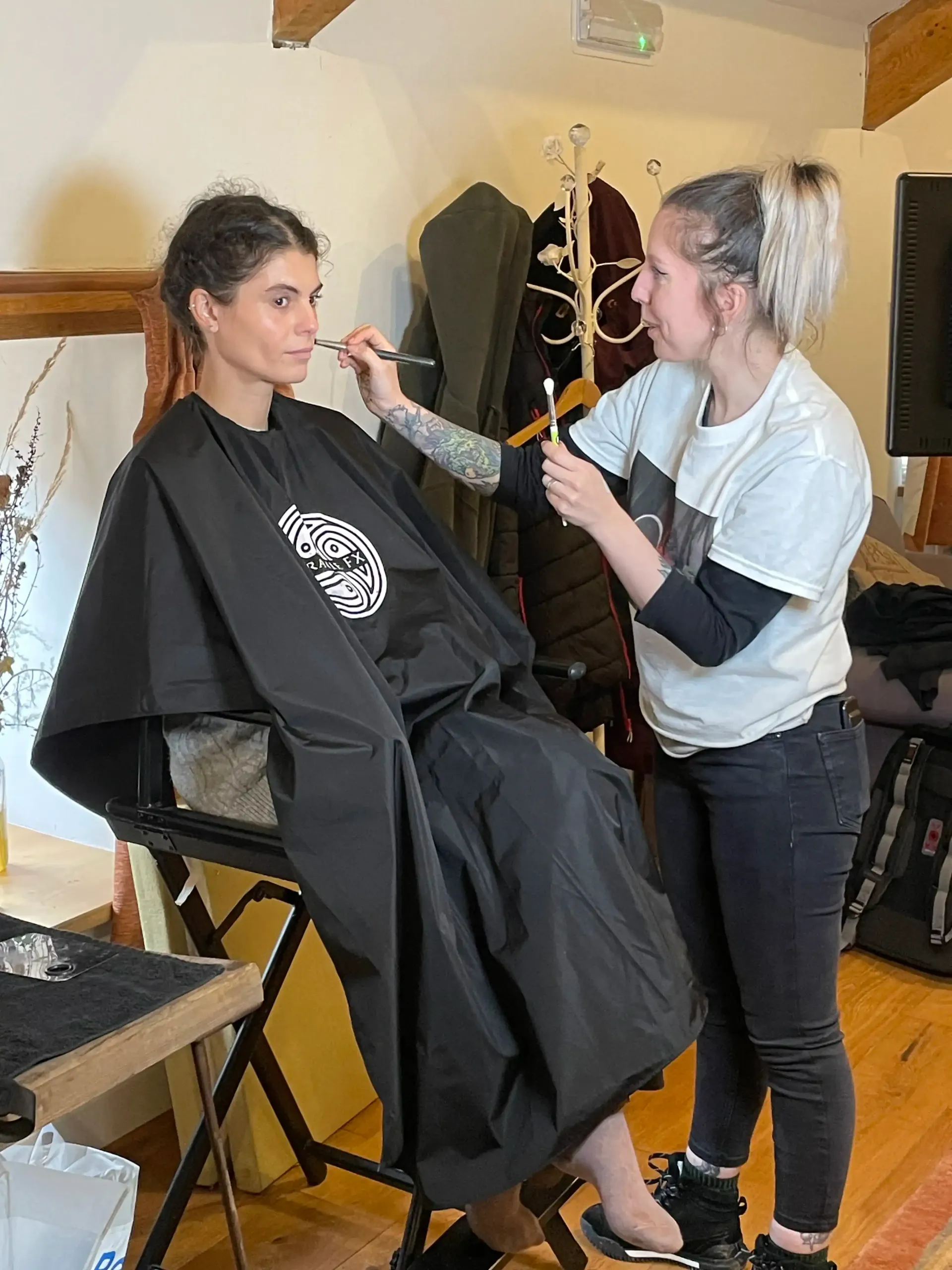 Person in a black cape getting makeup applied by another person in a room.