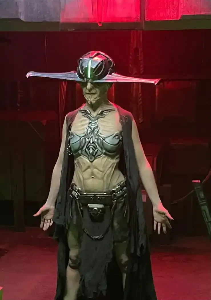 A person in a futuristic costume poses. They wear a headdress and body armor in a dimly lit setting.