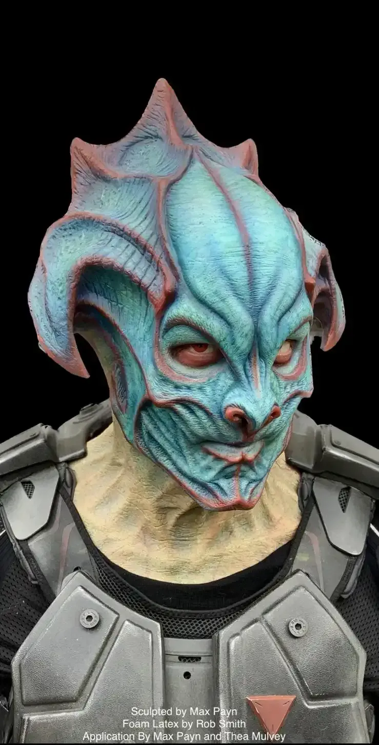 A person wearing a textured blue and red alien mask and grey tactical armor against a black background.