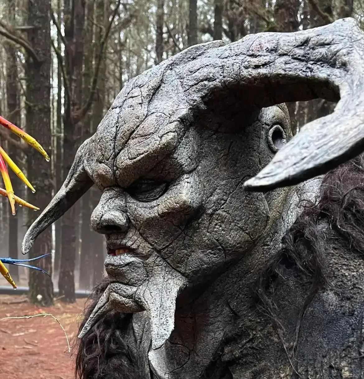 Close-up of a statue with large horns, stony face, and long beard. Forest background.