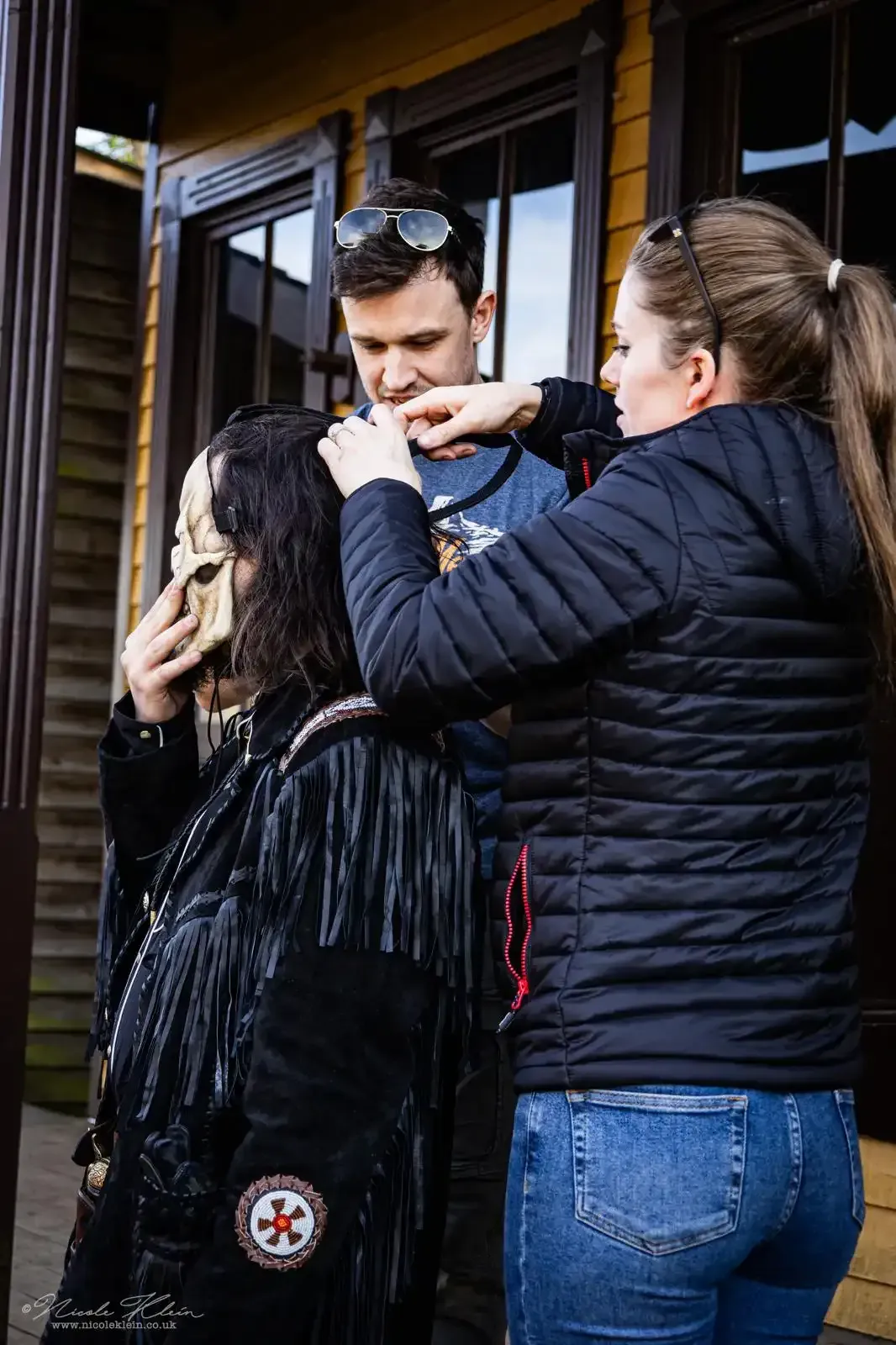 People dressed in costumes with a woman adjusting a mask, outdoors near a wooden building.