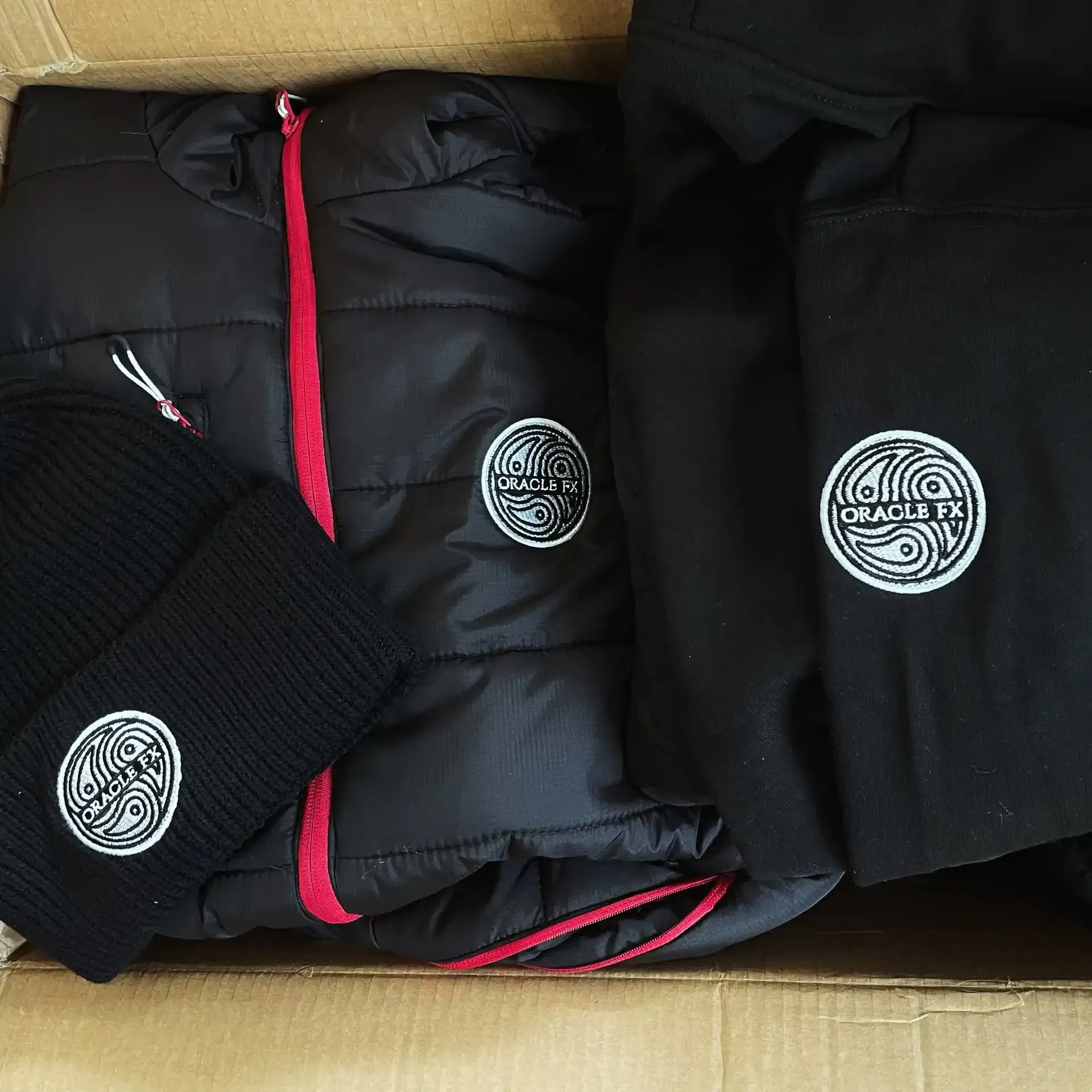 Black jacket, sweatshirt, and hat with logo in a cardboard box.