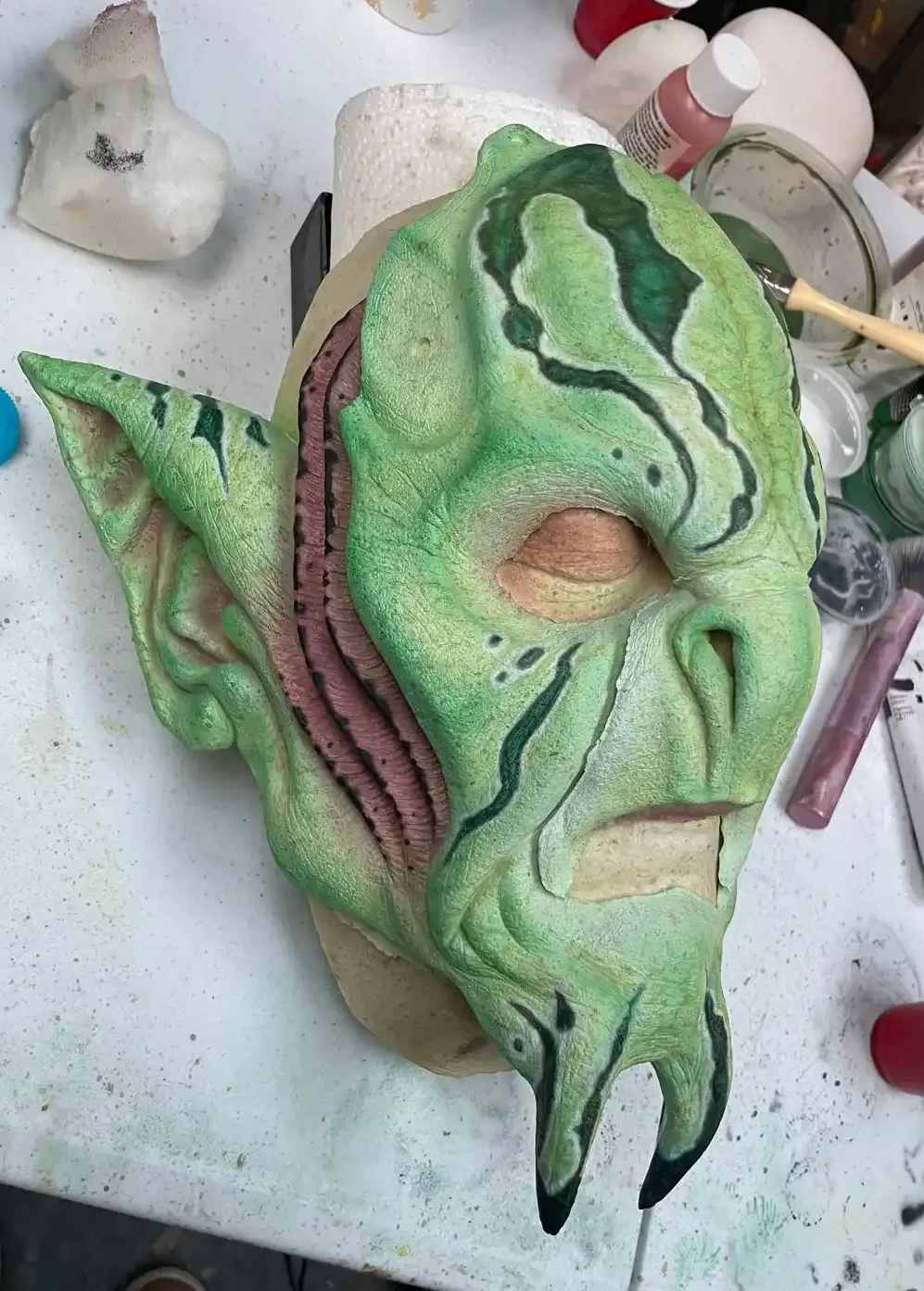 A detailed, green foam mask with pointed ears, textured facial features, and a beard, displayed on a workbench.