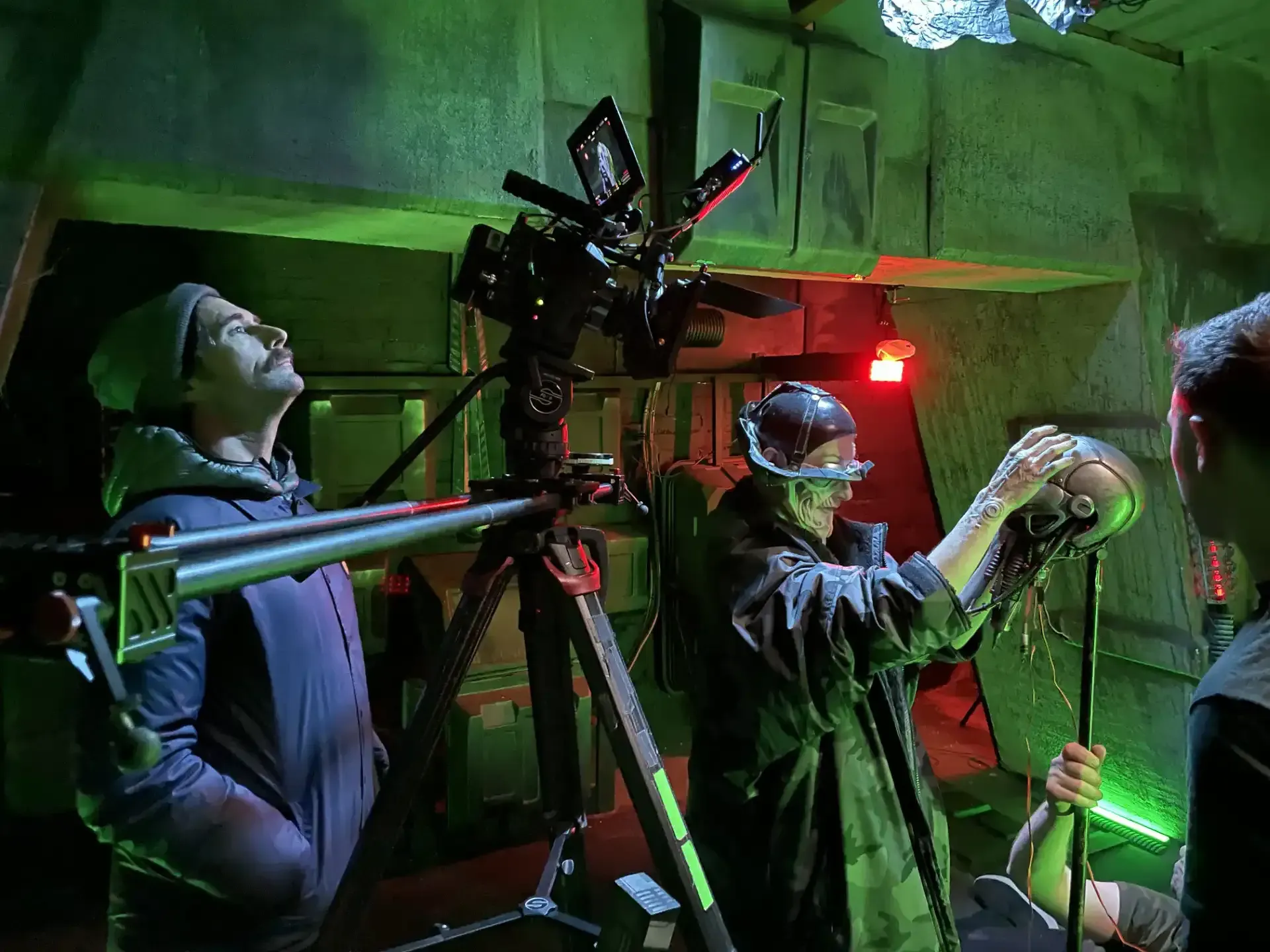A film set: Two people in costume work with equipment under green light. One looks up; the other holds a prop.