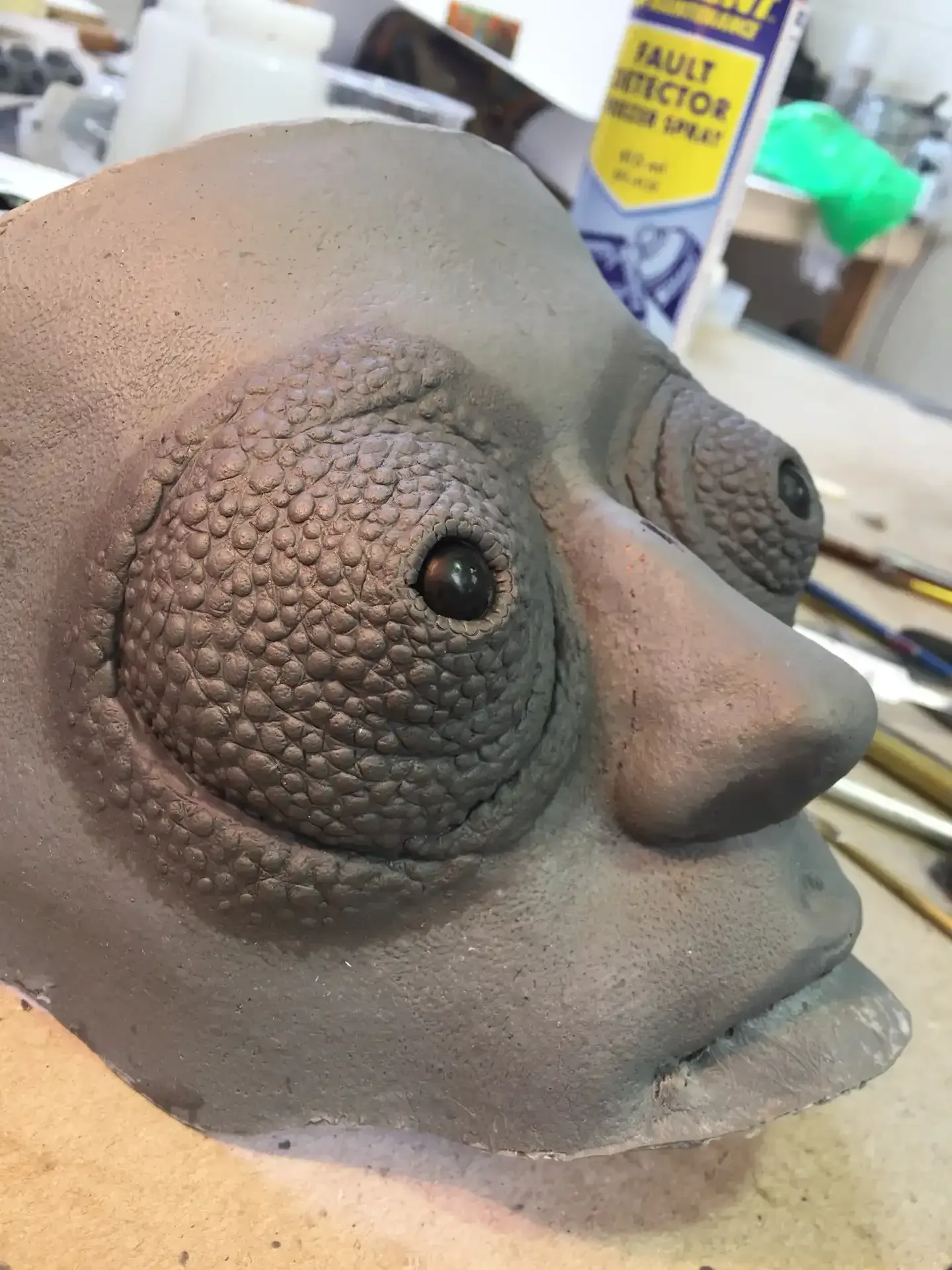 A gray, textured, alien-like clay mask sculpture with large bulging eyes, situated in an art workshop setting.