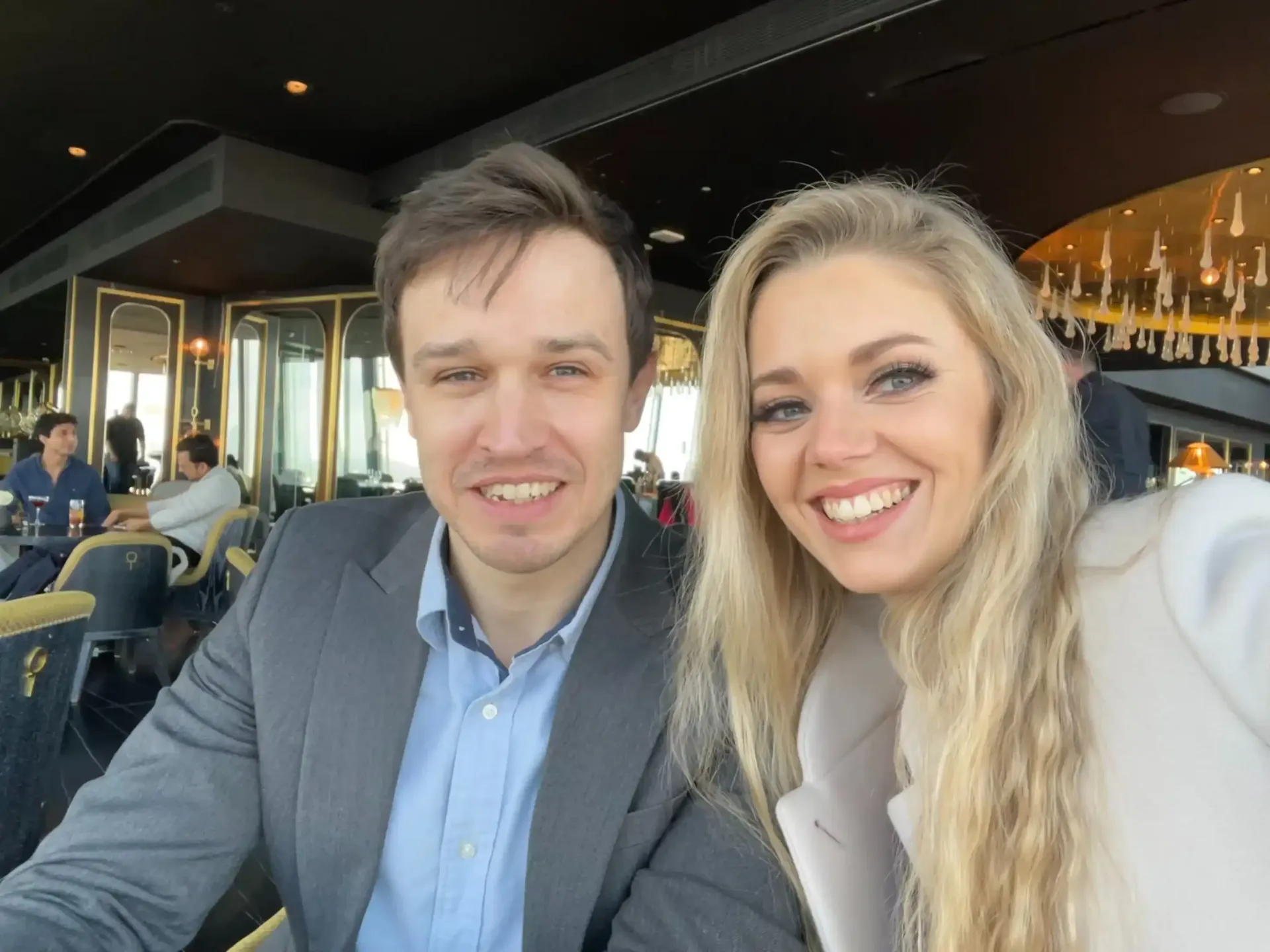 Man and woman smiling, taking a selfie in a restaurant with a panoramic view.