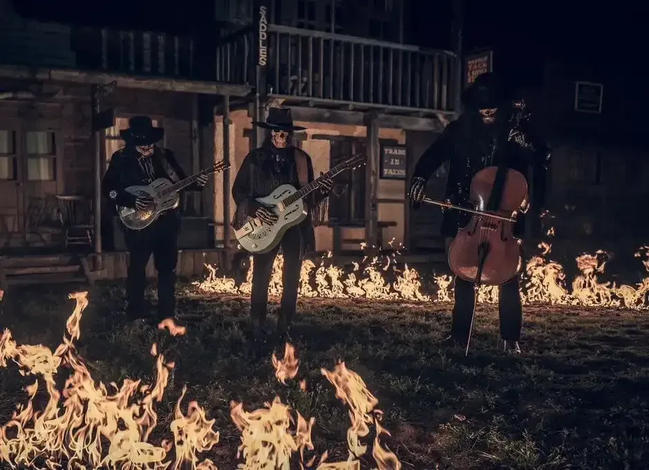 Three musicians in black suits playing instruments in front of a burning building at night.