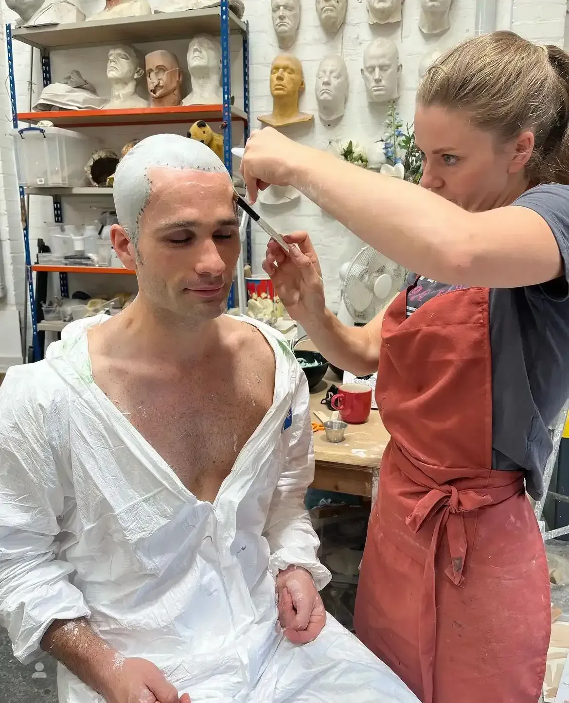 Person in white outfit has makeup applied by another with red apron, in workshop with masks.