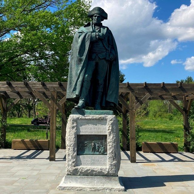 Bronze statue on stone pedestal in a park, with a pergola, trees, and blue sky behind it