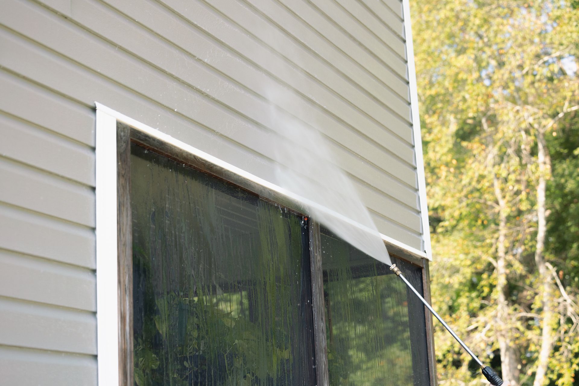 A person is using a high pressure washer to clean a window on the side of a house.
