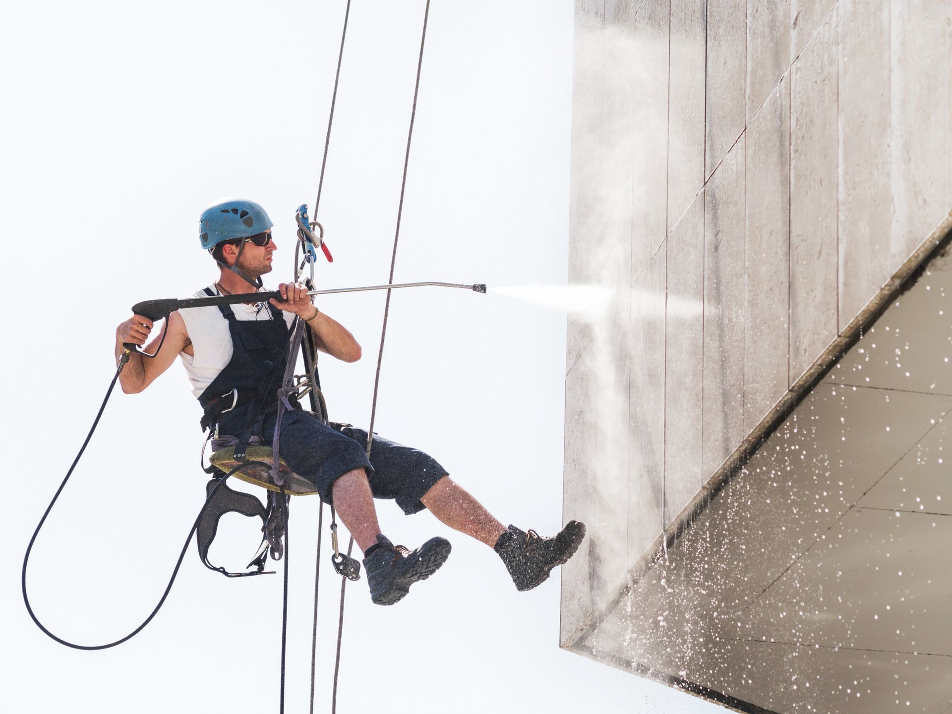 A man is sitting on a rope using a high pressure washer on a building.