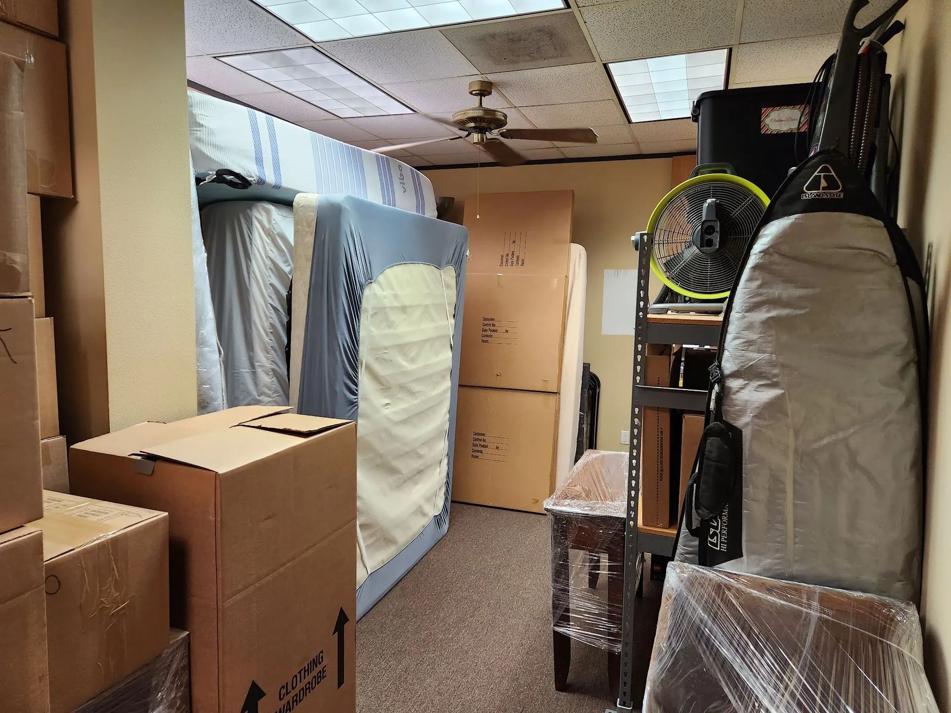 A narrow room filled with stacked cardboard boxes, mattresses, and various items wrapped in plastic for storage.