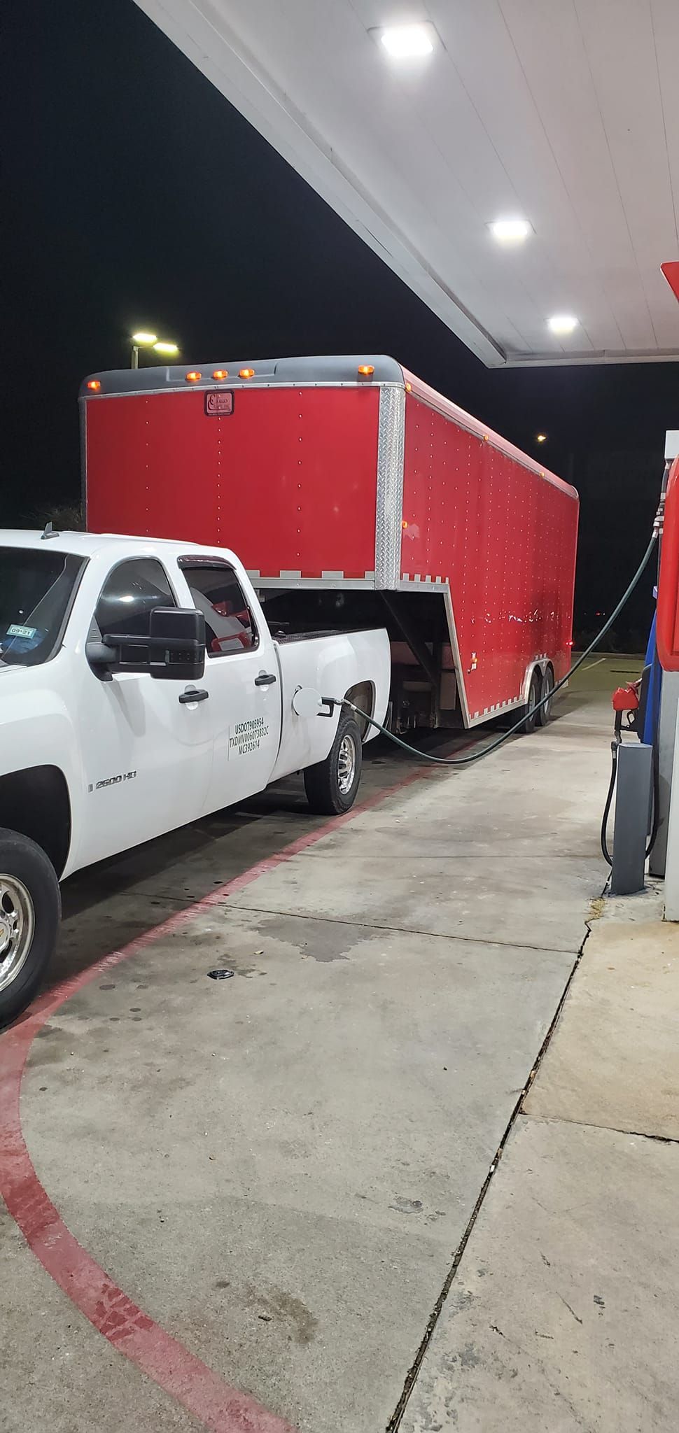 A white truck is towing a red trailer at a gas station.