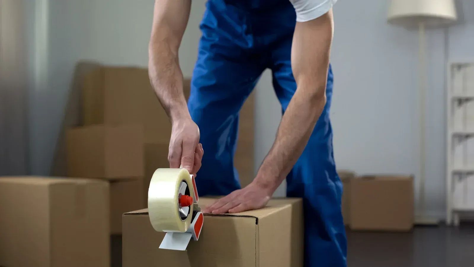 Person taping a cardboard box with packing tape in a room with stacked moving boxes.