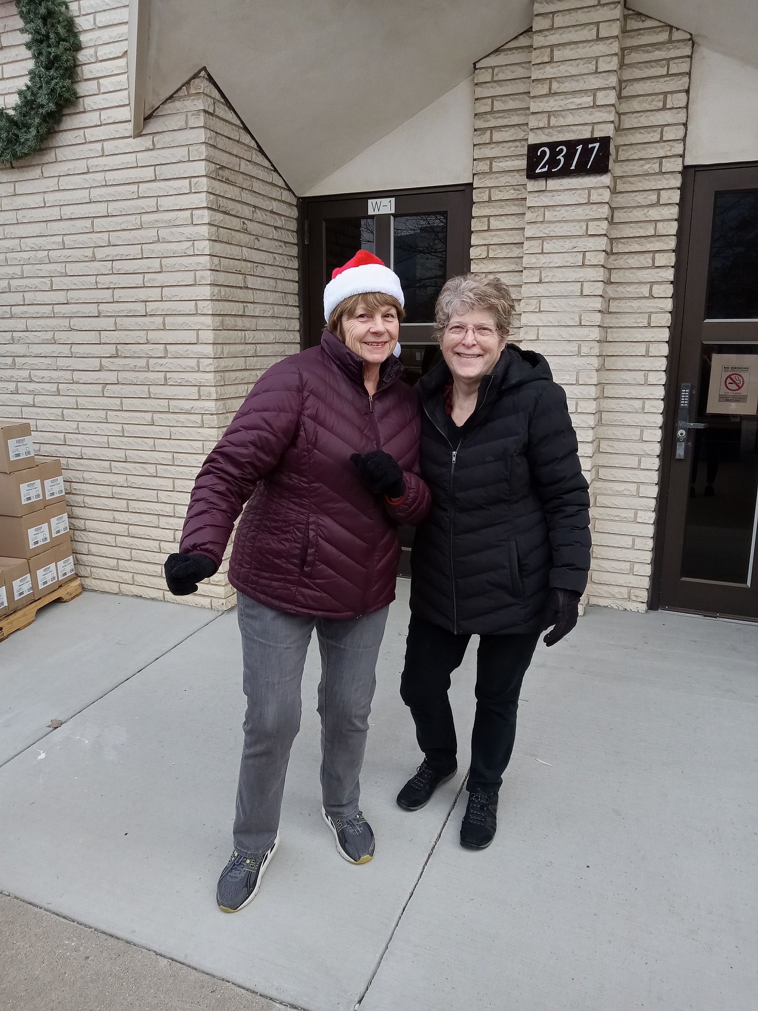 Two women wearing santa hats are posing for a picture in front of a brick building.