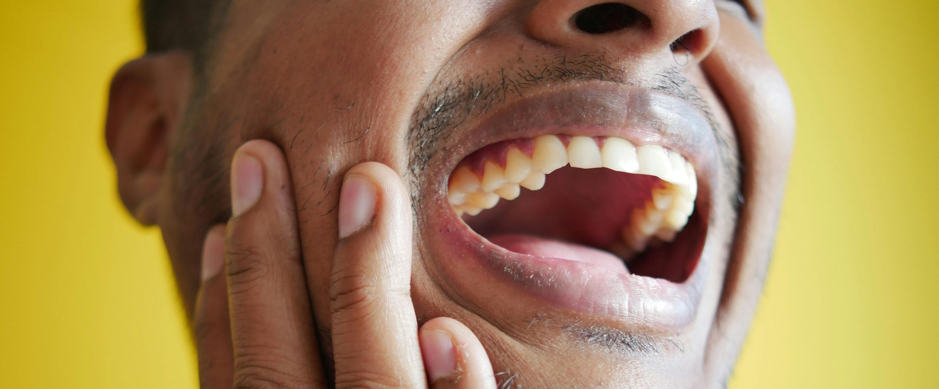 Close-up of a person's open mouth and face, hand touching the jaw. Yellow background.